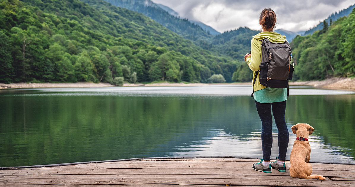 young woman standing on a dock with a dog. They are facing away from the camera looking out over some mountains. The woman is carrying a backpack. They look like they are about to set out on an adventure together.